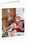 1970s Mother Giving First-Aid Putting Band-Aid On Knee Of Son In Kitchen by Anonymous