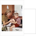 1970s Mother Giving First-Aid Putting Band-Aid On Knee Of Son In Kitchen by Anonymous