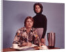 1970s Husband Wearing Loud Print Shirt Writing Family Budget and Wife Standing Behind Coffee Pot by Anonymous