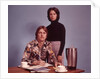 1970s Husband Wearing Loud Print Shirt Writing Family Budget and Wife Standing Behind Coffee Pot by Anonymous