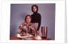 1970s Husband Wearing Loud Print Shirt Writing Family Budget and Wife Standing Behind Coffee Pot by Anonymous