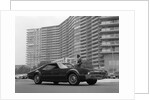 1960s Man In Suit and Hat Standing Beside Oldsmobile Toranado In Front Of Large Apartment Building by Anonymous