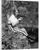 1920s Young Woman In Striped Skirt Sitting On Stream Bank In Woods Braiding Garland Of Wild Flowers by Anonymous