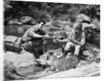Couple having picnic sitting on rocks by stream man pouring drink for woman from thermos by Anonymous