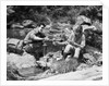Couple having picnic sitting on rocks by stream man pouring drink for woman from thermos by Anonymous