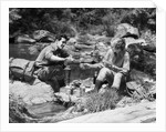 Couple having picnic sitting on rocks by stream man pouring drink for woman from thermos by Anonymous