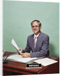 Smiling man at desk holding papers with mortgage officer sign looking at camera by Anonymous
