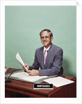 Smiling man at desk holding papers with mortgage officer sign looking at camera by Anonymous