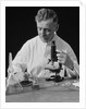 Male scientist wearing white lab coat writing data on clip board holding focus control of microscope by Anonymous