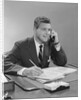 Businessman seated at desk talking on telephone smiling writing with pencil by Anonymous