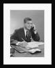 Businessman seated at desk talking on telephone smiling writing with pencil by Anonymous