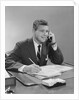 Businessman seated at desk talking on telephone smiling writing with pencil by Anonymous
