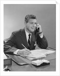 Businessman seated at desk talking on telephone smiling writing with pencil by Anonymous