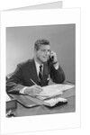 Businessman seated at desk talking on telephone smiling writing with pencil by Anonymous