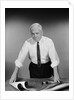 Middle aged man shirt sleeves rolled up standing blueprints on table by Anonymous