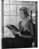 Elegant woman wearing pearls sitting by window reading book by Anonymous