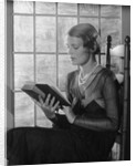 Elegant woman wearing pearls sitting by window reading book by Anonymous