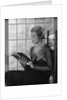 Elegant woman wearing pearls sitting by window reading book by Anonymous