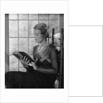 Elegant woman wearing pearls sitting by window reading book by Anonymous