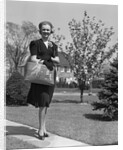 Stylishly dressed woman walking down suburban street carrying straw bag by Anonymous
