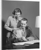 Couple man seated at desk holding paper woman standing looking over his shoulder smiling happy by Anonymous