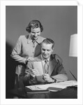 Couple man seated at desk holding paper woman standing looking over his shoulder smiling happy by Anonymous