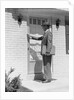 Salesman wearing suit tie hat carrying briefcase ringing suburban house doorbell by Anonymous