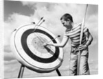 1960s boy jeans striped t-shirt holding bow & pulling arrow out of target bull's-eye by Anonymous