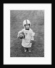 1950s boy standing in grass wearing oversized shirt & helmet holding football by Anonymous