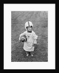 1950s boy standing in grass wearing oversized shirt & helmet holding football by Anonymous