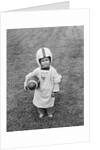 1950s boy standing in grass wearing oversized shirt & helmet holding football by Anonymous