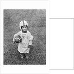 1950s boy standing in grass wearing oversized shirt & helmet holding football by Anonymous