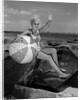 1960s smiling blond teenage girl sitting on rocky shore holding a beach ball waving looking at camera by Anonymous
