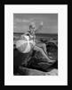 1960s smiling blond teenage girl sitting on rocky shore holding a beach ball waving looking at camera by Anonymous