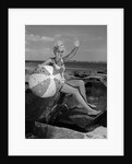 1960s smiling blond teenage girl sitting on rocky shore holding a beach ball waving looking at camera by Anonymous