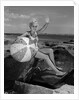 1960s smiling blond teenage girl sitting on rocky shore holding a beach ball waving looking at camera by Anonymous