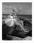 1960s smiling blond teenage girl sitting on rocky shore holding a beach ball waving looking at camera by Anonymous