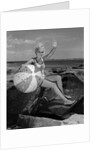 1960s smiling blond teenage girl sitting on rocky shore holding a beach ball waving looking at camera by Anonymous