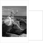 1960s smiling blond teenage girl sitting on rocky shore holding a beach ball waving looking at camera by Anonymous