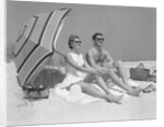 1960s couple in sunglasses sitting on beach blanket with legs extended with umbrella by Anonymous