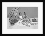 1960s couple in sunglasses sitting on beach blanket with legs extended with umbrella by Anonymous