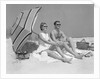 1960s couple in sunglasses sitting on beach blanket with legs extended with umbrella by Anonymous