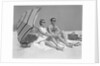 1960s couple in sunglasses sitting on beach blanket with legs extended with umbrella by Anonymous
