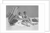 1960s couple in sunglasses sitting on beach blanket with legs extended with umbrella by Anonymous