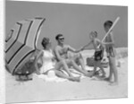 1960s at the beach two boys with baseball bat trying to pull up father seated next to mother so he will play with them by Anonymous
