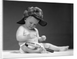 1960s baby in diaper wearing sherlock holmes deerstalker style double-billed cap with magnifying glass resting on leg inspecting a clue by Anonymous