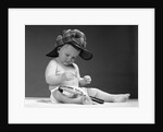 1960s baby in diaper wearing sherlock holmes deerstalker style double-billed cap with magnifying glass resting on leg inspecting a clue by Anonymous