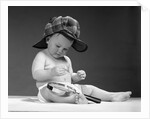 1960s baby in diaper wearing sherlock holmes deerstalker style double-billed cap with magnifying glass resting on leg inspecting a clue by Anonymous