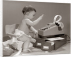 1960s baby seated in small chair hitting keys on office adding machine on top of small file drawers by Anonymous