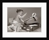 1960s baby seated in small chair hitting keys on office adding machine on top of small file drawers by Anonymous